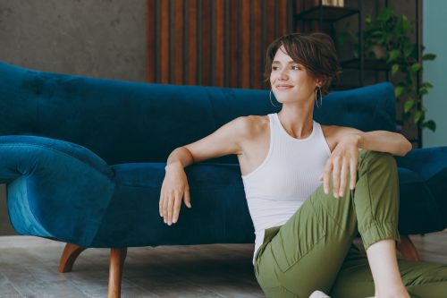 woman with short hair wearing green pants and a white tank top sitting on the floor in front of a blue velvet couch