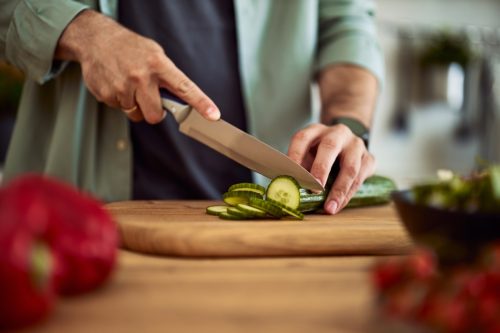 A close-up shot of male hands using a sharp kitchen knife to cut a fresh cucumber for a salad on a wooden cutting board.