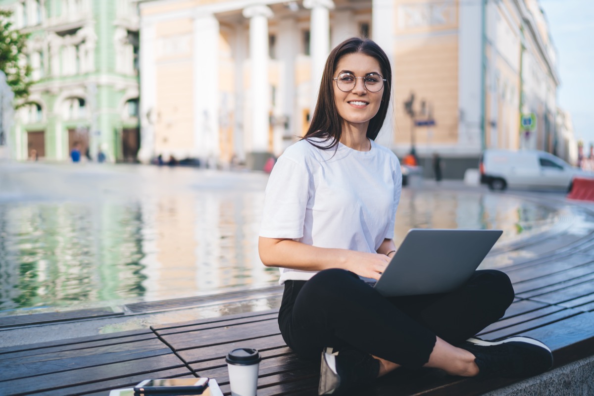 Happy digital nomad with modern netbook sitting at city urbanity and thinking about idea for web projecting during freelance working, carefree Caucasian hipster girl with laptop smiling outdoors