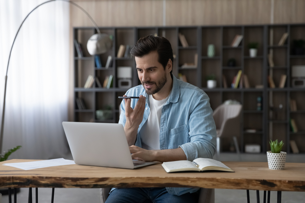 A man sitting on his computer while talking to his phone's virtual assistant