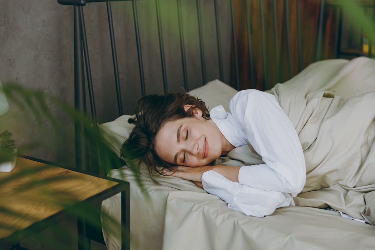 Young calm woman wear white shirt pajama she lying in bed hands folded under head rest relax spend time in cosy bedroom lounge home in own room hotel wake up dream be lost in reverie good mood day.