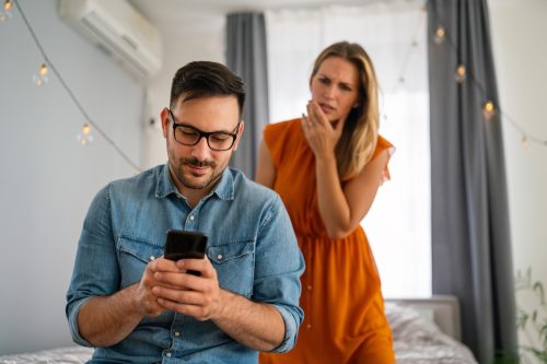 Jealous woman looking on as man uses cell phone while sitting