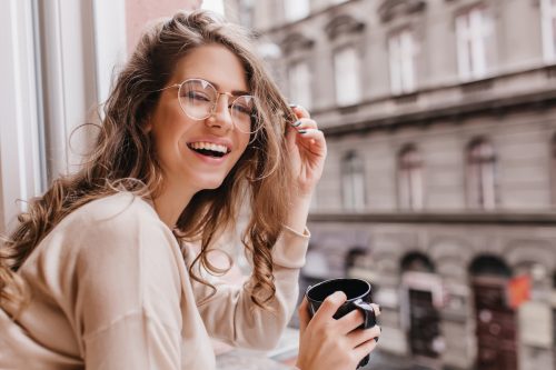 Close-up portrait of laughing brunette girl in beige sweater drinking coffee on city background.