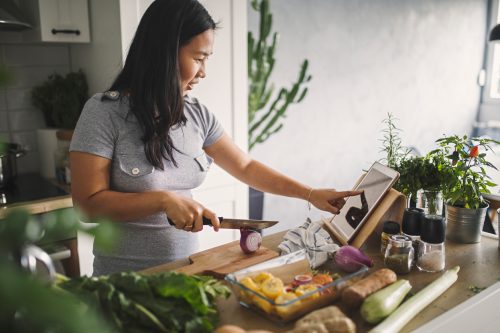 A young woman cooking in her kitchen while reading a recipe off of her tablet