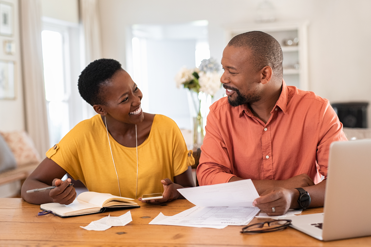 Middle-aged couple sitting at a table in their home paying bills while smiling.