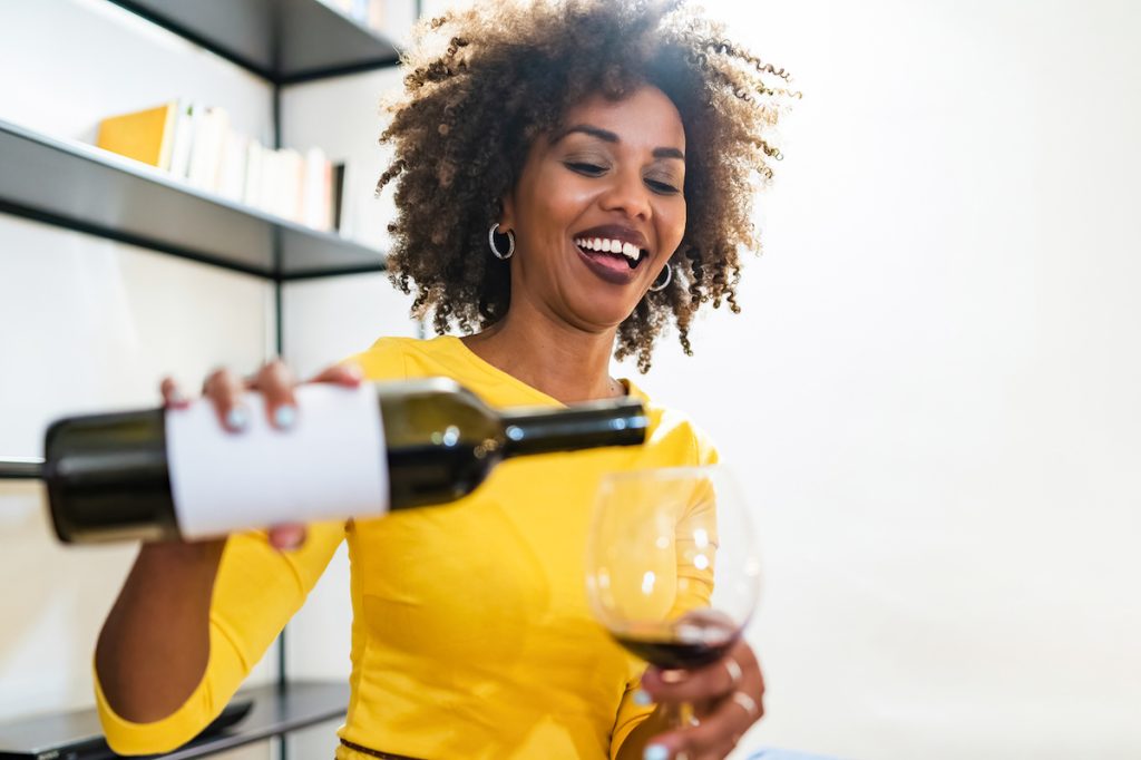 A smiling woman wearing a bright yellow shirt pours herself a glass of red wine.