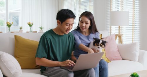 young family man woman smile happy sit at home sofa couch pay for hotel booking, search choose, buy ticket on laptop social media app travel trip getaway plan tour with cute small pet dog.