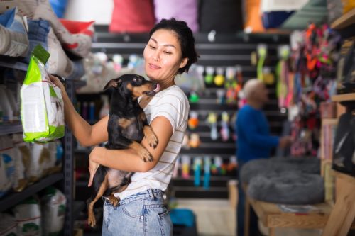 woman with little dog in hands standing in salesroom of pet shop and choosing dog food.