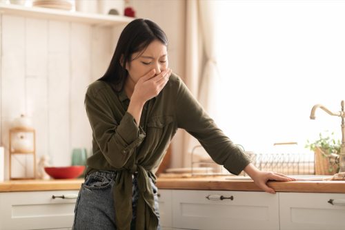 Young woman in casual feeling nausea, cover mouth, standing next to table in kitchen, copy space. Toxicosis during pregnancy, food poisoning concept