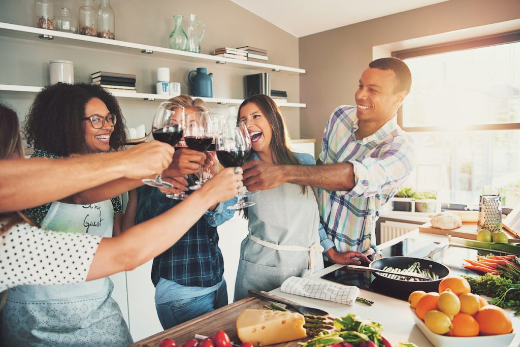 Group of young cheerful friends toasting with wine glasses while cooking in kitchen.