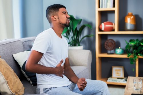 Shot of a young man suffering from stomach pain while lying sitting a sofa at home