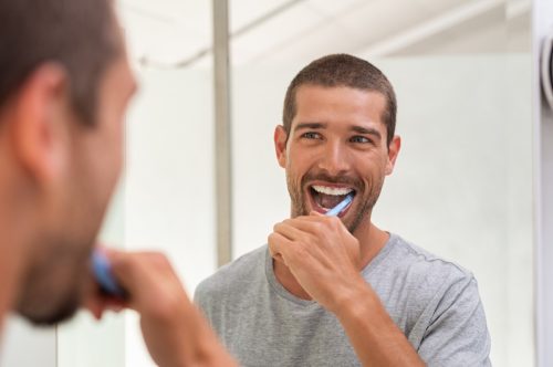 Smiling young man with toothbrush cleaning teeth and looking mirror in the bathroom. Handsome man brushing his teeth in morning in bathroom. Happy guy in pajamas brushing teeth before going to sleep.