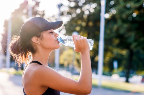 Closeup of a sporty-looking young woman wearing a black baseball cap drinking from a water bottle outside