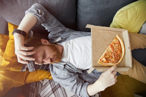 Ảnh chụp từ trên xuống chân dung một người đàn ông có râu đang nằm trên ghế dài và ăn pizza trong khi xem TV ở nhà, không gian sao chép