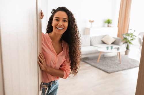 Welcome. Portrait of cheerful woman standing in doorway of modern apartment, greeting visitor and inviting guest to enter her home, happy smiling young lady holding door looking out flat