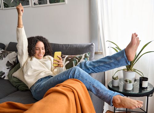 Happy excited young latin woman relaxing on couch using phone winning money in online app game. Young lucky woman feeling winner looking at cellphone, receiving great news or discount offer.