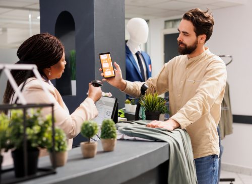 African American woman cashier scanning Black Friday mobile promo code from customer smartphone, using barcode scanner. Young man shopper using coupon to get discount while shopping in clothing store