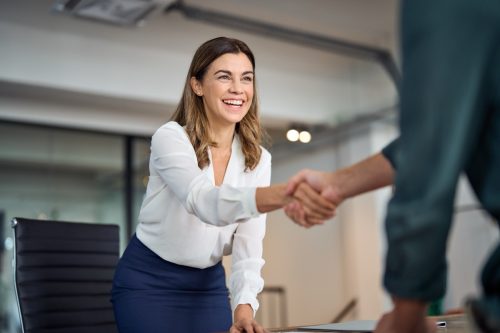 Happy mid aged business woman manager handshaking greeting client in office. Smiling female executive making successful deal with partner shaking hand at work standing at meeting table