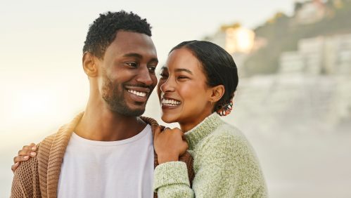 Fun, free and happy couple laughing and hugging at the beach, enjoying fresh air and time together. Young lovers talking and bonding while being affectionate, sharing a funny joke while walking