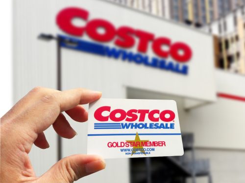 Cận cảnh một bàn tay đang cầm thẻ thành viên Costco với một cửa hàng Costco ở phía sau