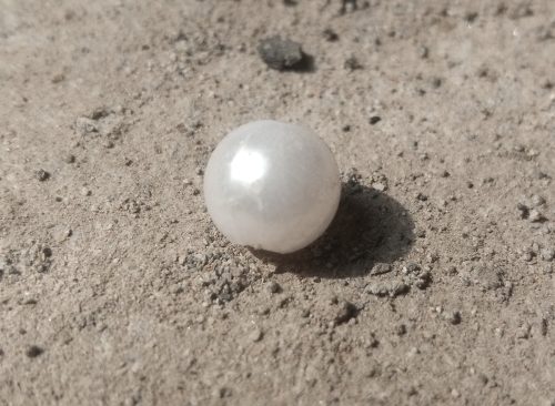 A close-up of a single, white pearl nestled in the coarse grains of light brown sand. The pearl is smooth and round with a faint iridescence. The background is blurred.