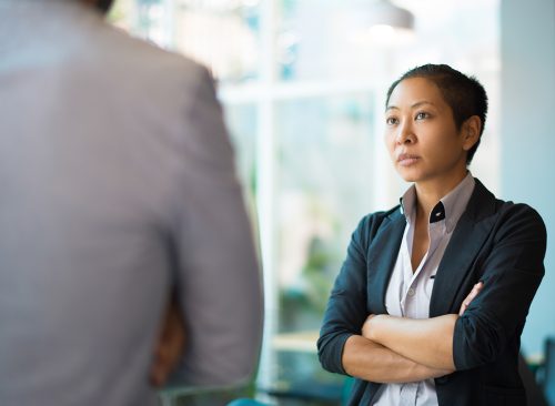 Tense Asian businesswoman looking at male partner with crossed arms. Two colleagues confronting each other in office space. Clashing personalities concept