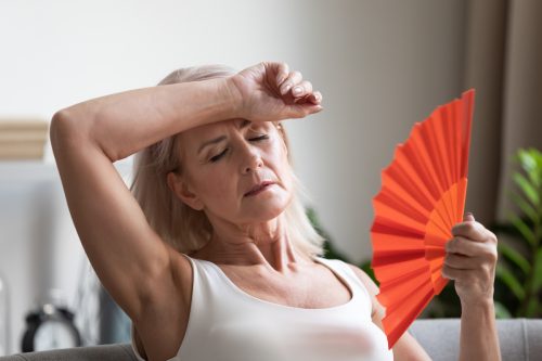 A tired-looking senior woman sitting on her couch fanning herself on a hot day